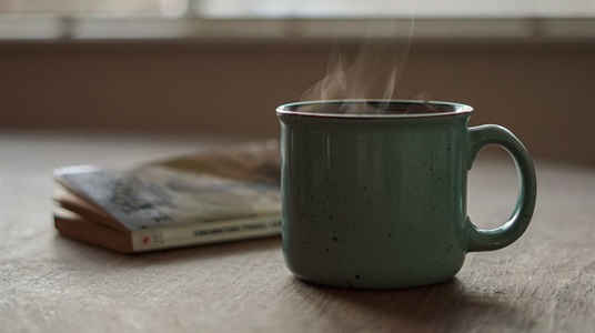 A mug with steam rising from it rests next to a paperbook book.