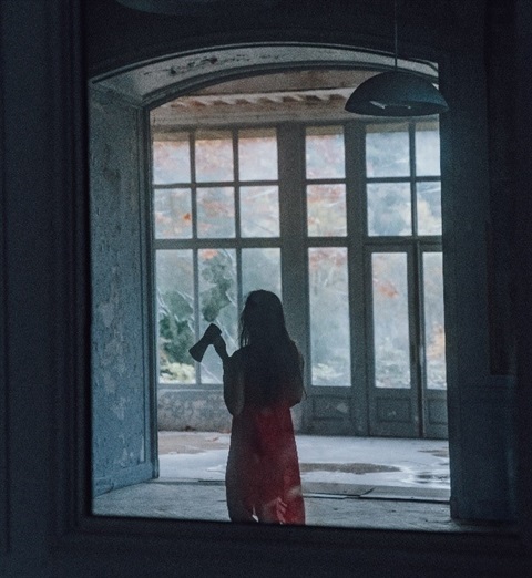 A long-haired woman holding an axe is seen in silhouette, while she stands in front of the interior windows of a large and grand room. 