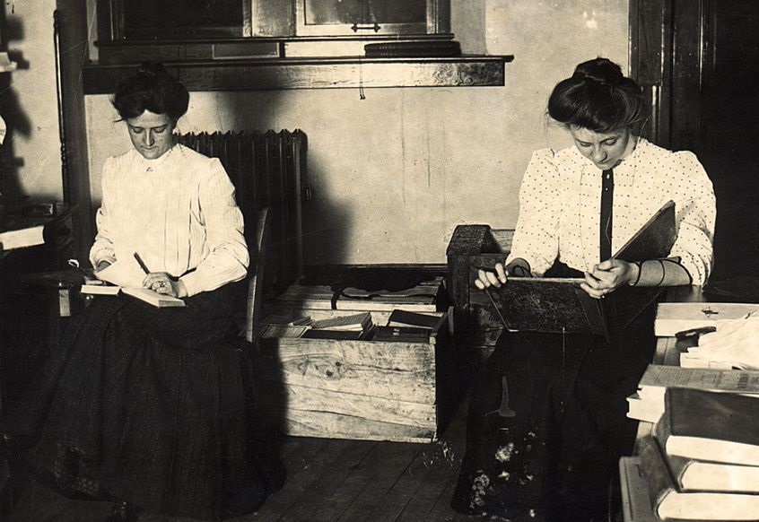 Two early twentieth century libraries sit doing paperwork at the Carnegie library